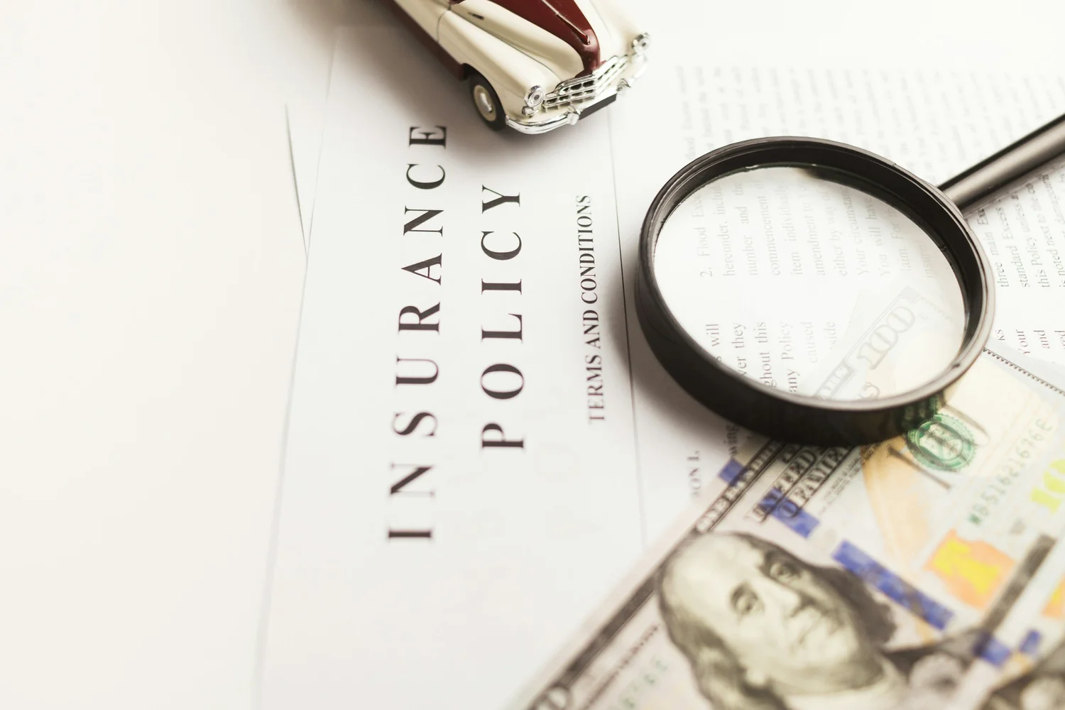 Insurance policy document with a magnifying glass, hundred-dollar bills and a toy car