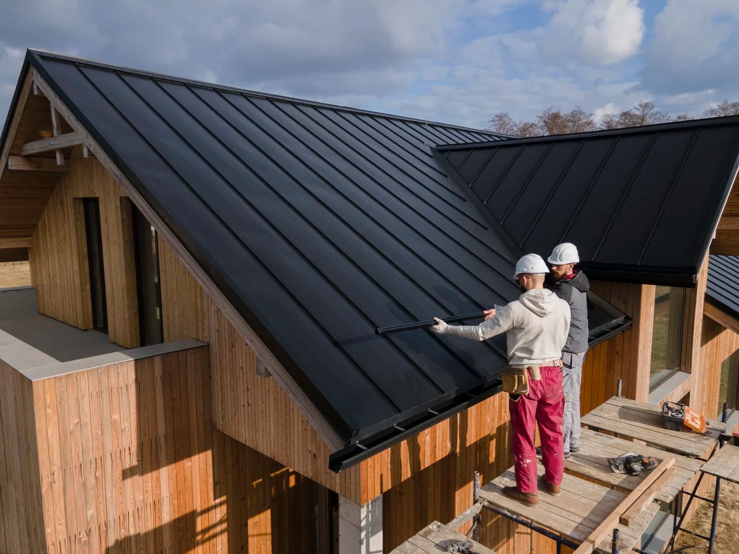 Two workers in white hard hats on scaffolding beside a black standing seam metal roof