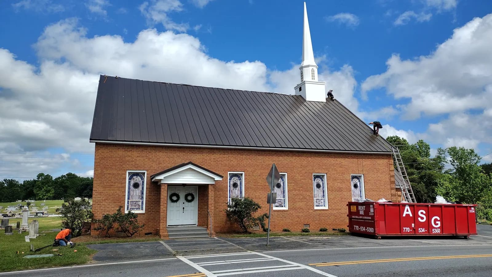 Crew working on a new dark standing seam metal roof on a brick church, with the job's dumpster at the curb