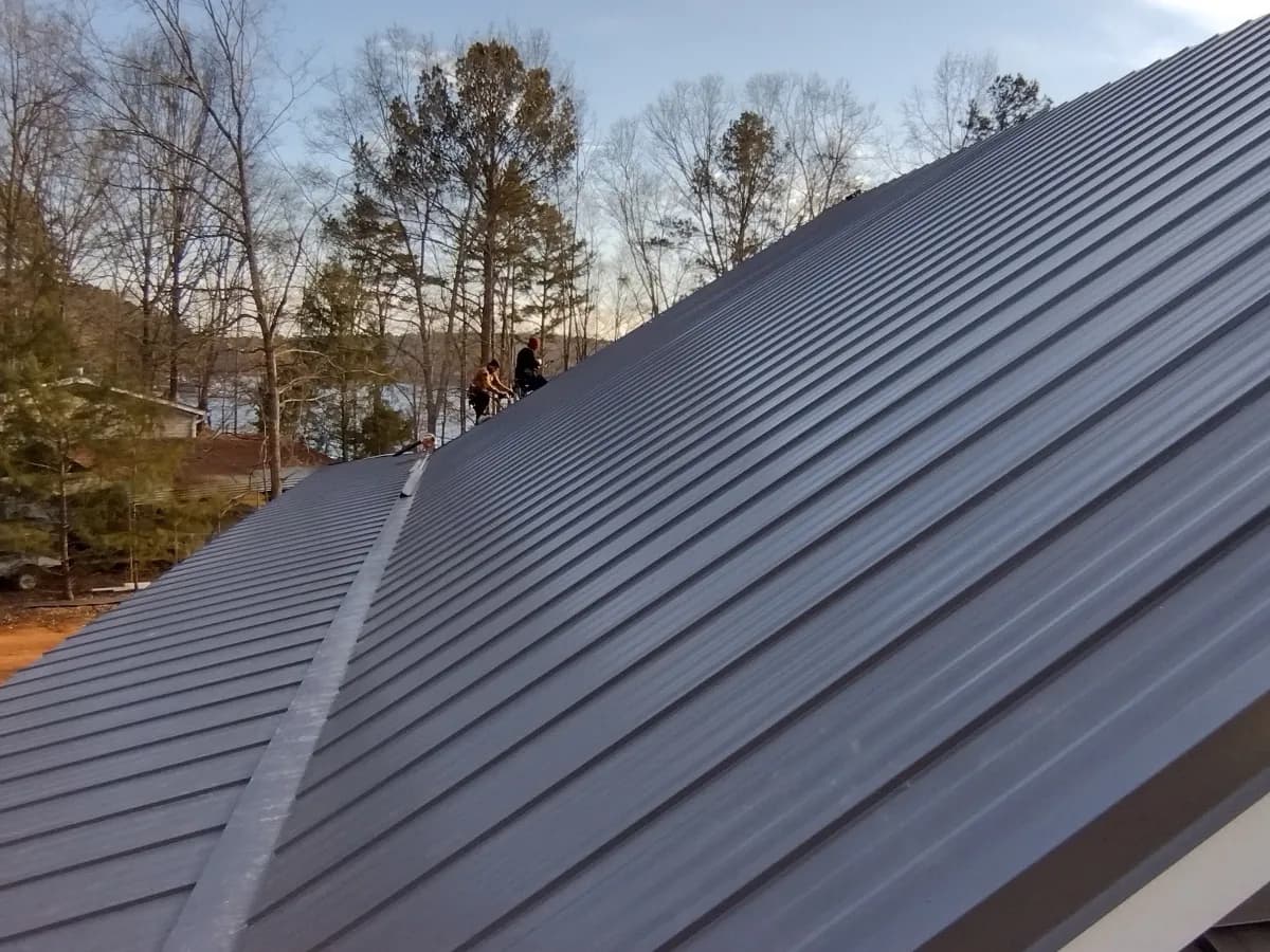 Two roofers near the ridge of a new charcoal standing seam metal roof, with a lake behind the trees