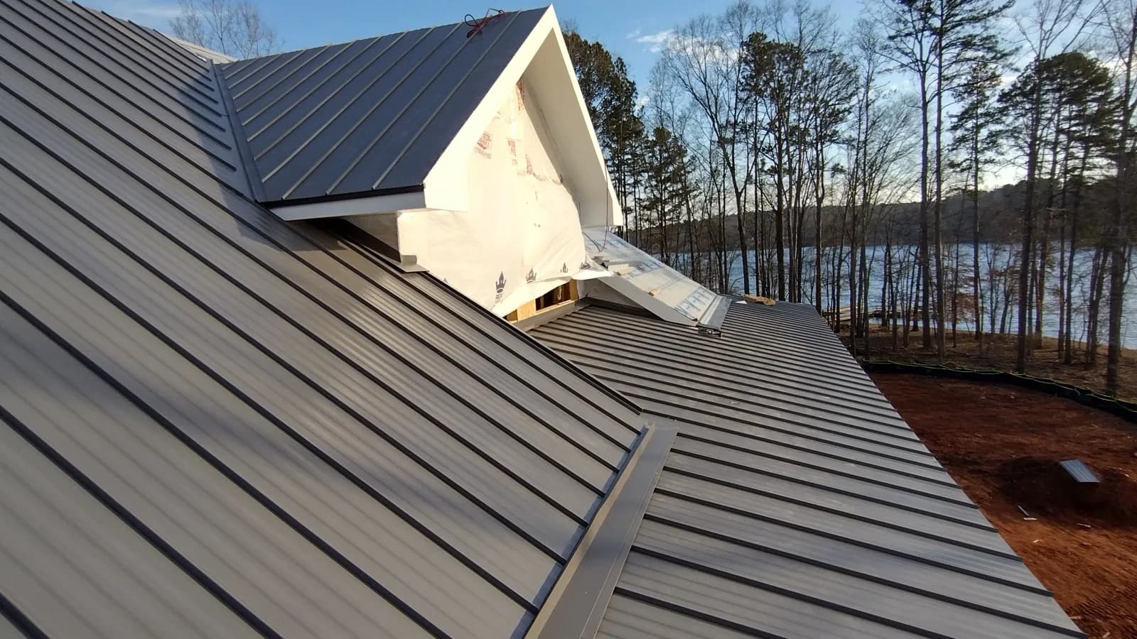 Charcoal standing seam metal roof meeting a dormer gable, with a lake through the trees