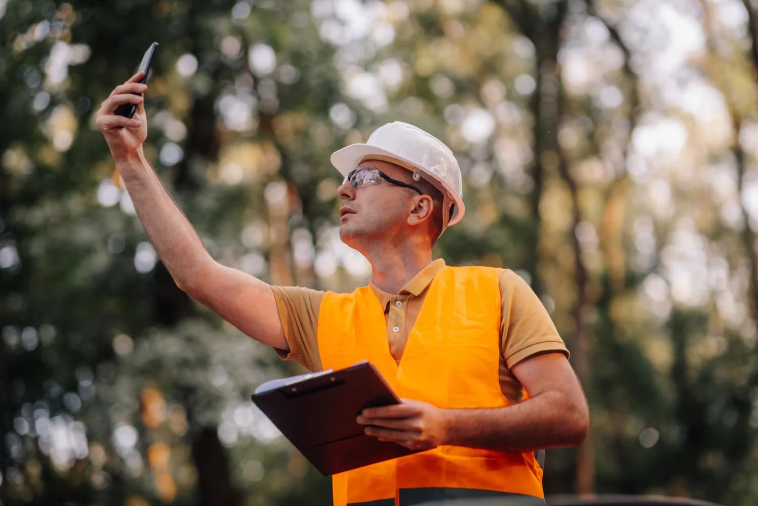 Man in a white hard hat and orange safety vest holding up a phone and a clipboard