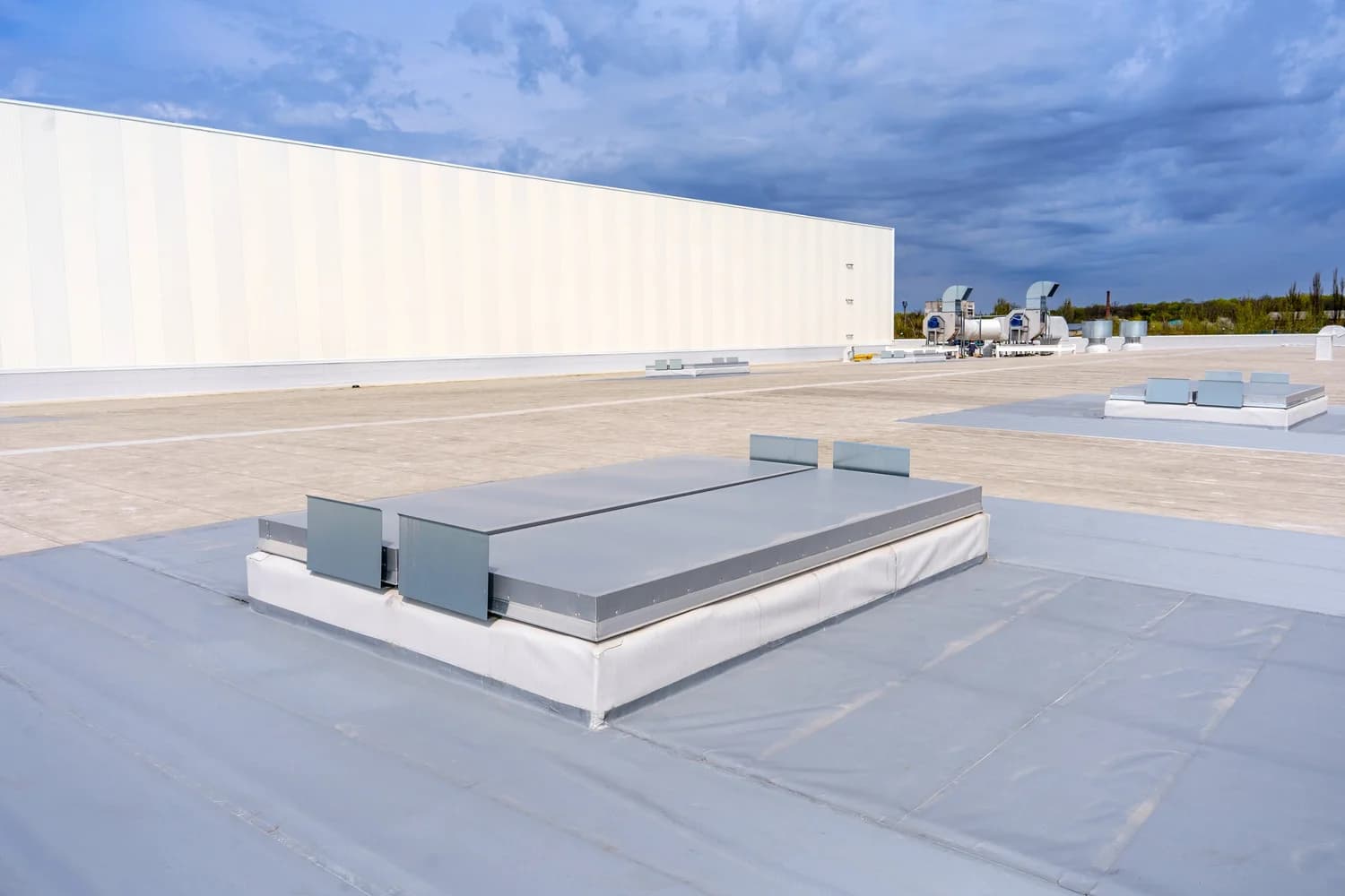 Flat commercial roof with a gray membrane, rooftop hatches and ventilation units beside a white wall