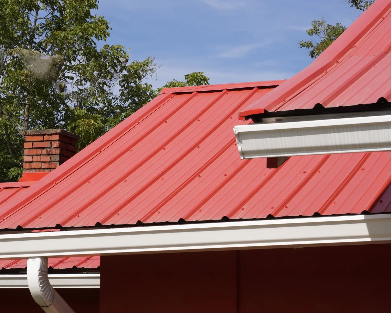 Red exposed-fastener metal roof with white gutters, a downspout and a brick chimney