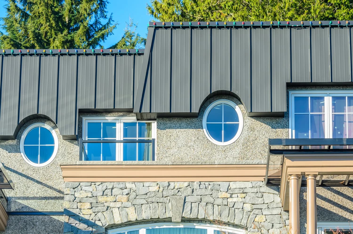 Dark gray standing seam metal roof over round windows and a stone and stucco facade