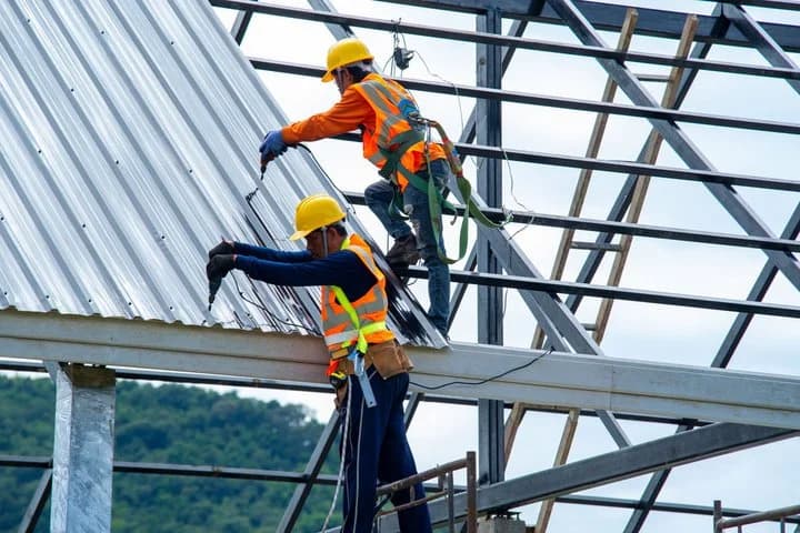Two workers in hard hats and safety harnesses fastening metal roof panels to a steel frame