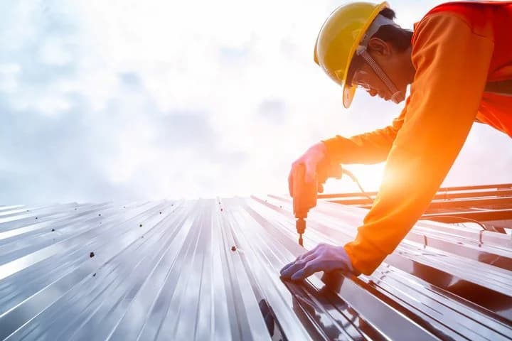 Worker in a yellow hard hat driving screws into ribbed metal roof panels with a drill