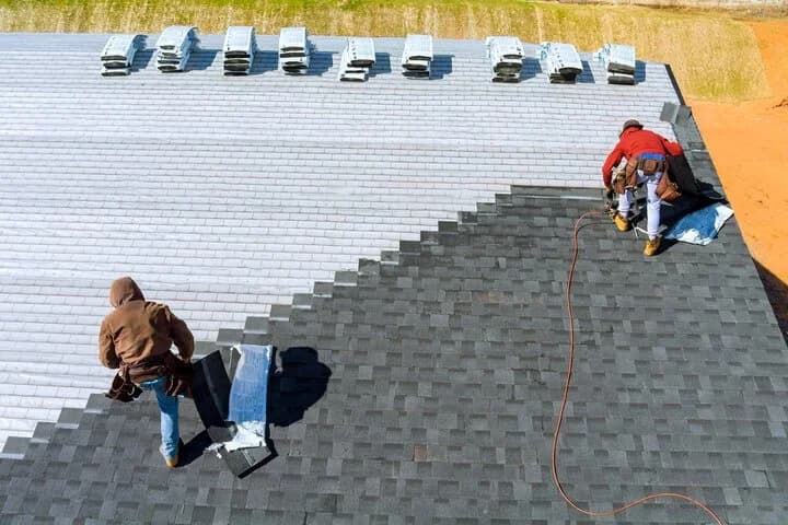 Two roofers nailing dark gray shingles over white underlayment, with bundles stacked along the ridge