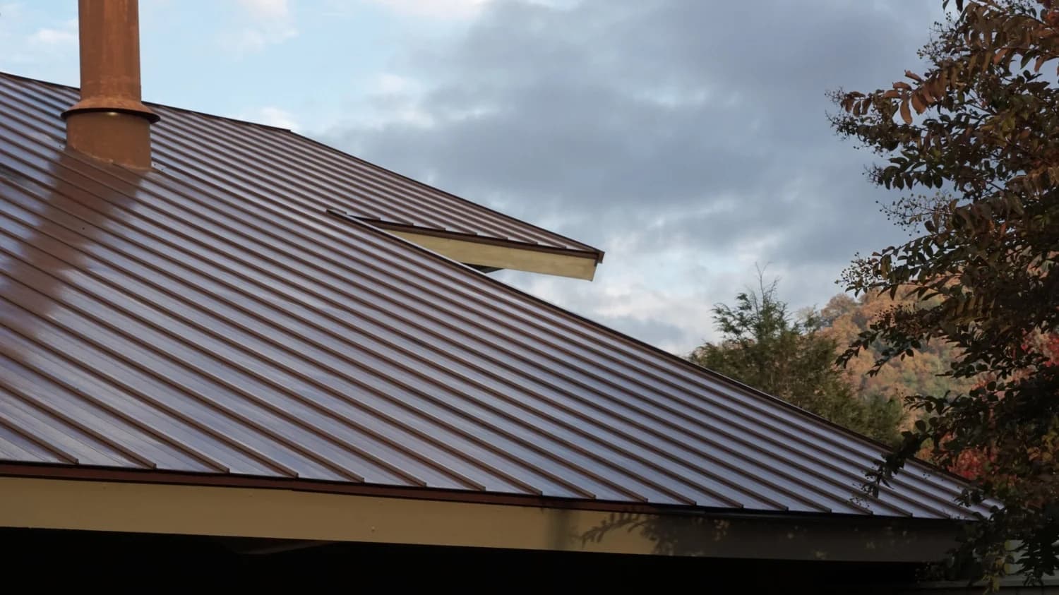 Brown standing seam metal roof with a rust-colored flue pipe under a cloudy sky
