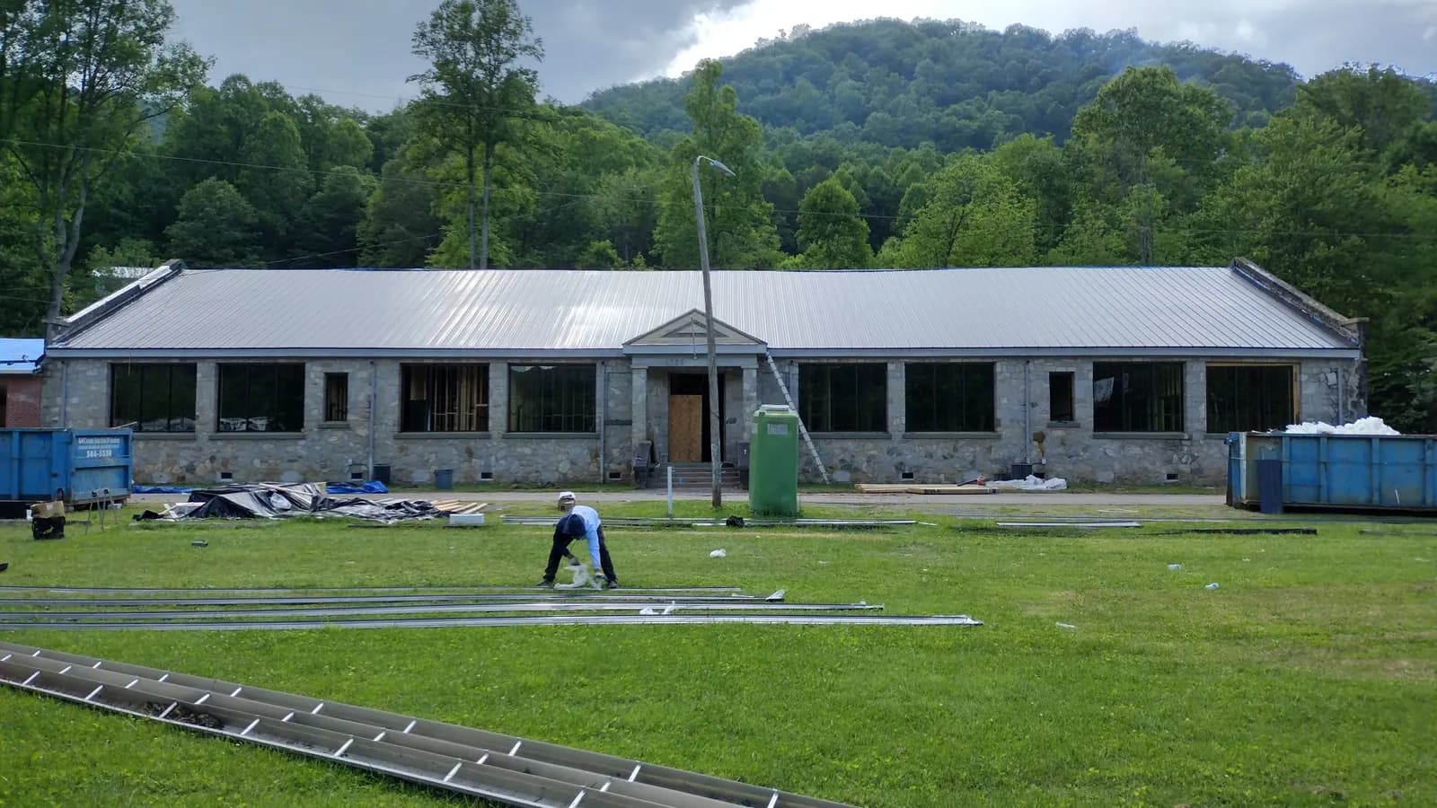 Crew cutting metal panels beside a stone building getting a new metal roof, with wooded mountains behind